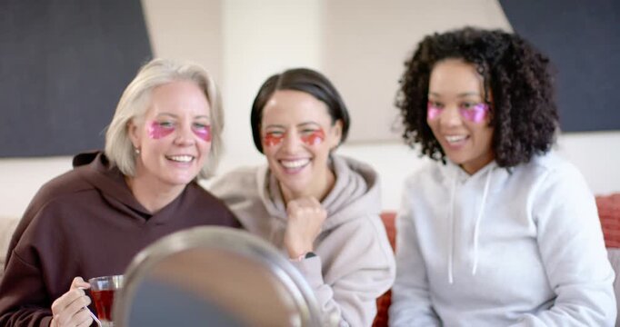 Diverse female friends leaning to mirror, laughing and adjusting under-eye patches for skincare