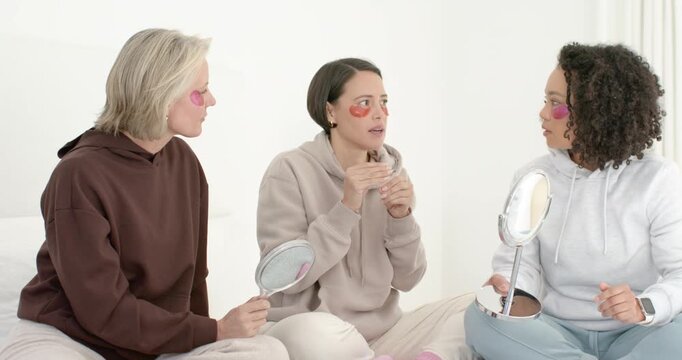 Three women in hoodies sitting on bed, center woman opening jar passing mirrors testing patches