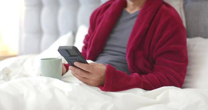 Senior Asian man receiving phone message, reading christmas message and smiling in bed with mug