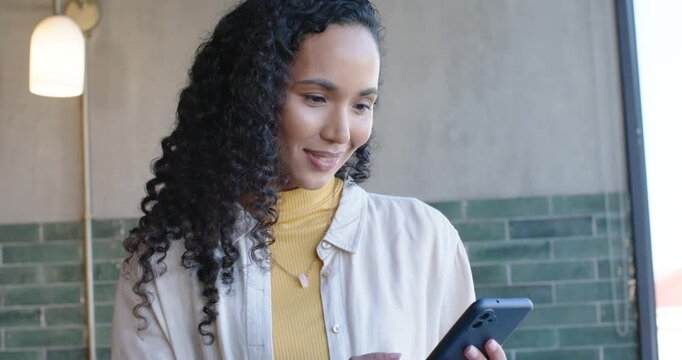Woman wearing mustard top, reading phone text, tapping right thumb, smiling in cafe