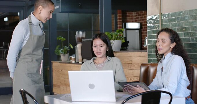 Diverse female coworkers at cafe table with laptop, tablet, gesturing to server in apron speaking