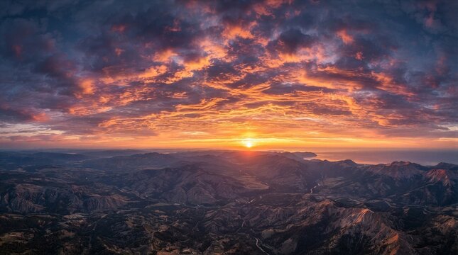 Sun dips low over rugged mountain range. Sky glows with fiery orange and purple hues. Clouds stretch wide, catching sunset's final light. Valley sprawls below, bathed in warm golden glow