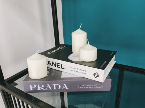 Belarus, Svetlogorsk, September 12, 2026. Fashion Books with White Candles. Stack of Chanel and Prada books topped with three white candles on a glass shelf. Elegant interior decor.