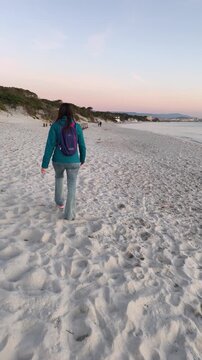 Vertical slow motion rear view of a woman walking barefoot on the sand at sunset in winter, with warm evening light and a peaceful coastal atmosphere.