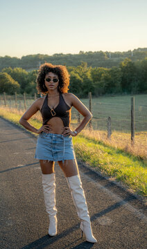 Black woman standing on quiet rural road during warm sunset light, wearing sunglasses, black halter top, denim skirt, and white boots, with hands on hips against peaceful countryside fields.