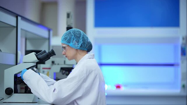Wide side view of a female scientist in a blue hair net adjusting a microscope stage and looking through the eyepieces, with cold blue light glowing in a modern lab