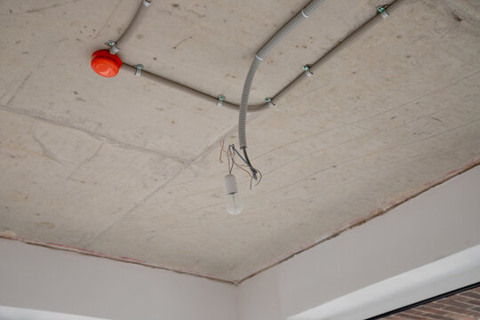 Open electrical wiring on concrete ceiling with cables in corrugated conduit and temporary bulb socket. Red junction box mounted on slab. Renovation and installation work.