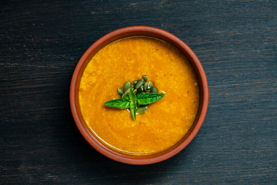 Pumpkin cream soup with seeds and basil top view, close up. Minimalist flat lay of hot orange pumpkin or carrot cream soup garnished with seeds and green leaves in clay bowl on dark wooden background
