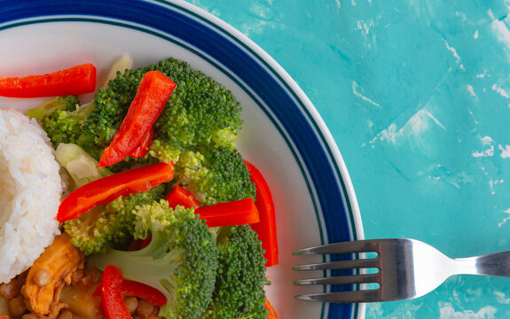 Fresh broccoli and red peppers sit beside white rice. Fork rests beside the plate, ready to eat. Bright turquoise background adds a clean, lively feel. The meal looks healthy and appetizing, vibrant