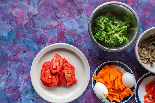 Vibrant vegetables arranged on a textured purple background. Fresh broccoli, carrots, tomatoes, and mushrooms add color. Eggs and sliced red peppers complete the healthy meal setup