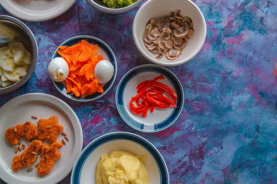 Colorful bowls hold fresh, vibrant ingredients. Mushrooms, peppers, carrots, eggs, and more. Preparation begins on a vivid, textured background. Food arrangement suggests a healthy, creative meal