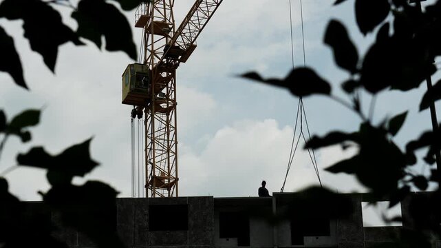 Construction worker standing atop high rise building guides heavy concrete slab suspended from tower crane. Builder maneuvers large precast panel carefully near structural edge during daytime.