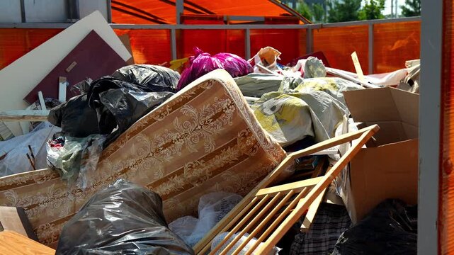 Discarded mattress sits atop pile of junk in municipal dumpster outdoors under bright sunlight. Cluttered heap of broken household furniture and plastic garbage bags fills refuse container.