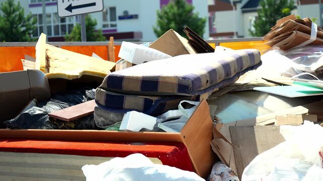 Discarded domestic items and broken furniture gather inside large garbage dumpster near residential area. Messy assortment includes wooden debris, foam mattress, and discarded household clutter.