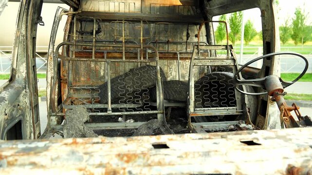 Burnt out bus cabin stands completely destroyed outdoors showing rusted metal frames exposed seat springs and charred steering wheel while vehicle remains motionless beside tree lined road.