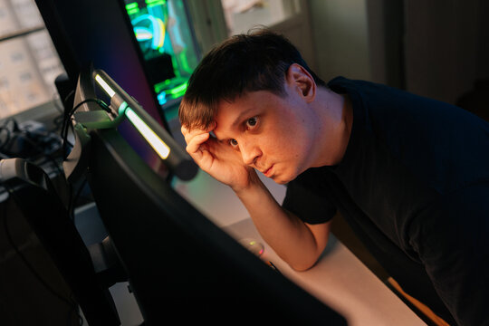 Concentrated young man with thoughtful expression works or plays on computer at night. Angled shot shows intense focus under monitor light. Modern workspace with neon glow creates moody atmosphere.