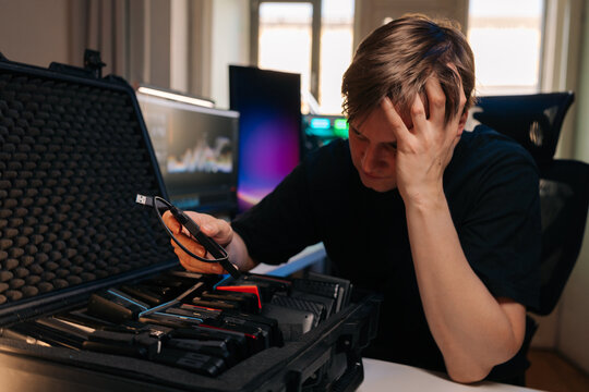 Worried young man sitting at desk with technical issue. Frustrated professional holding head, looking at many external hard drives in protective case. Concept of data loss, backup failure, IT problem.
