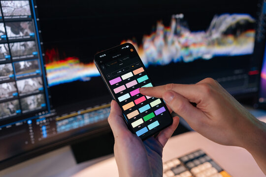 Hands use smartphone app selecting colors for video editing. Monitor with post production software and waveform in background. Modern digital workflow, filmmaking, color grading concept.
