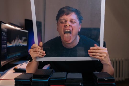 Young man at desk making humorous expression with tongue out and face pressed against clear glass panel, creating playful and silly moment in home office setting.