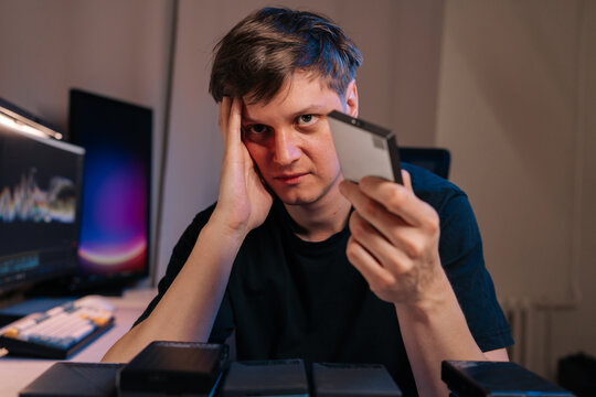 Man feeling overwhelmed and stressed while holding external hard drive, surrounded by other storage devices and computer, struggling with technology and data management issues.