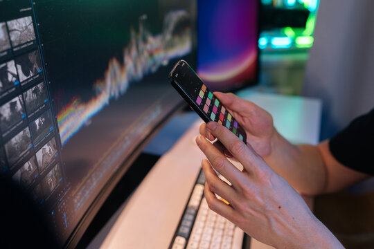 Person hands holding smartphone displaying color palette application, using it for detailed color correction while editing video footage on curved monitor in dimly lit workspace.