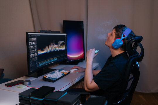 Young man wearing headphones and using vape pen, blowing smoke while focused on editing software displayed crossdual monitor workstation setup.