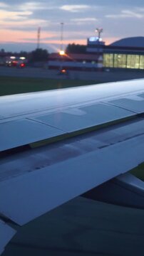 Low-angle passenger window view shows airplane wing with partially extended flap as aircraft taxis beside illuminated terminal at dusk, with airplane, wing, flap, aircraft detail, showing jet