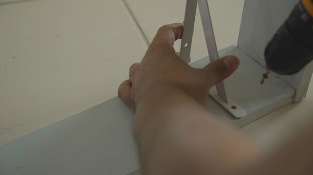 Close up of hand using an electric drill driver to fix a white bracket on a wooden shelf