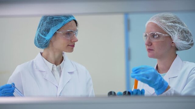 Two female scientists in hair nets and safety glasses examine test tubes of orange liquid and a dropper at the lab bench, front view framed through laboratory shelving