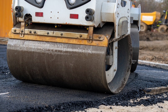 Heavy machinery compacts asphalt during road construction near a building site