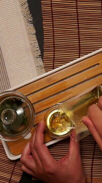 Top view close up of pouring freshly brewed yellow tea from glass pitcher vessel into small glass cup on wooden tea tray during traditional oriental tea ritual.