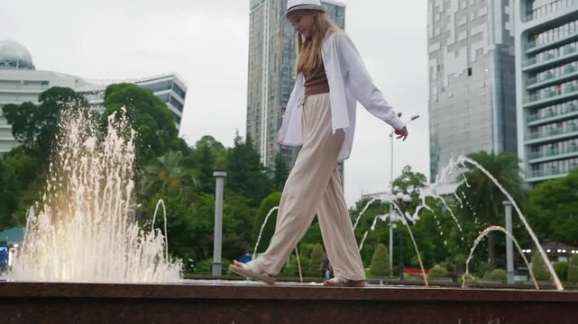 Woman in white coat, beige pants, gray hat balances on fountain ledge in modern city plaza with tall buildings, low angle camera captures confident movement over water jets and urban architecture.