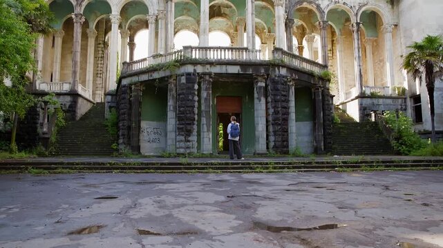 Single person stands near deteriorating neoclassical building with grand colonnade, moss covered stone, overgrown vegetation, static wide shot capturing architectural decay, urban exploration theme
