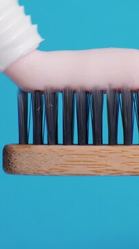 Squeezing pink toothpaste smoothly over dark bristle rows of eco friendly toothbrush. Toothbrush holds thick layer of paste against vivid blue backdrop for daily dental health maintenance.