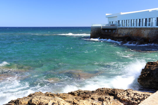 Long exposure in Nea Chora, Chania, Crete. Silky smooth turquoise waves crash against the rocky Mediterranean coastline under a clear summer sky.