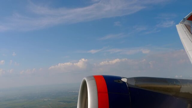 Passenger airplane turning in flight, jet engine and wing seen through window above green farmland and clouds, sunny day, handheld view