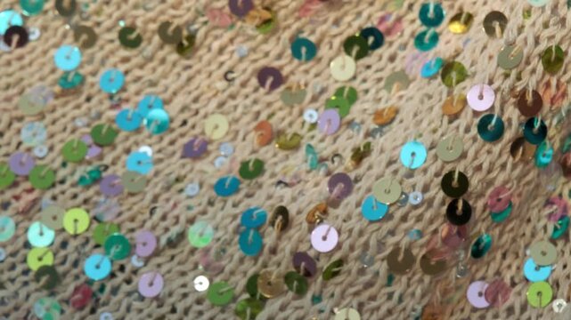 A close up video capturing colorful sequins and their reflections shimmering on a light brown handmade knitted textile texture, highlighting intricate details and glittering effects.
