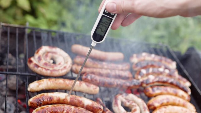 Hand inserts digital food thermometer probe into sizzling sausage on outdoor charcoal grill. Cook monitors internal temperature rise on electronic display screen during barbecue cooking.