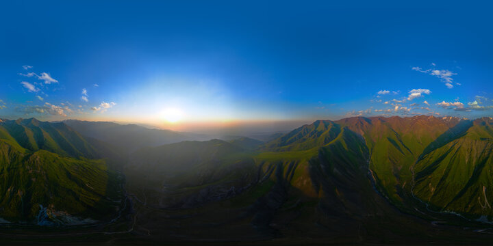 Full spherical panoramic view of a mountain landscape in Kyrgyzstan. Green slopes and valleys are illuminated by warm light from the low sun, creating a hazy atmosphere.
