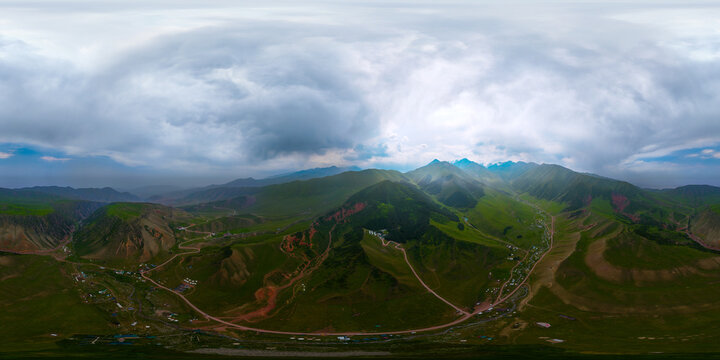 360-degree aerial panorama captures a mountainous landscape with green hills and rocky inclines. A dirt road connects small settlements and tents, under a partly cloudy sky.