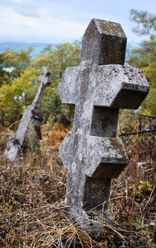 Vertical view of an old leaning stone cross in an overgrown cemetery. Low angle view of a weathered gravestone in an abandoned graveyard. Historic christian monument against a cloudy sky