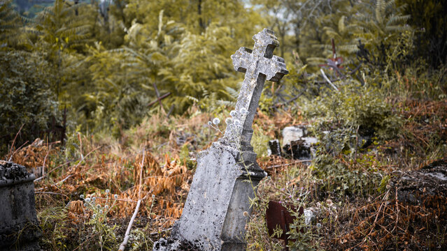 Old leaning stone cross gravestone in an overgrown cemetery. Weathered abandoned tombstone surrounded by dry grass and weeds in autumn. Spooky historical graveyard background
