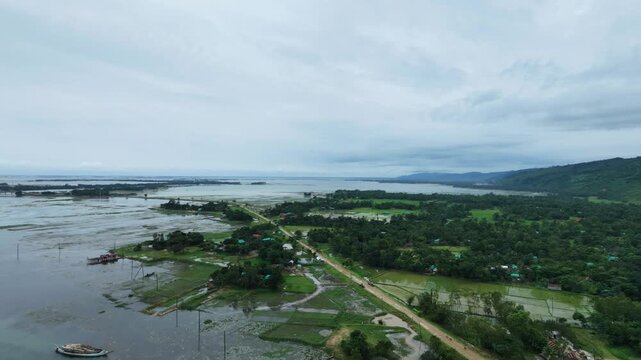 The drone sweeps across the long country dirt road splitting the flooded green fields from the main river bank, with the Indian mountainside on the right.
