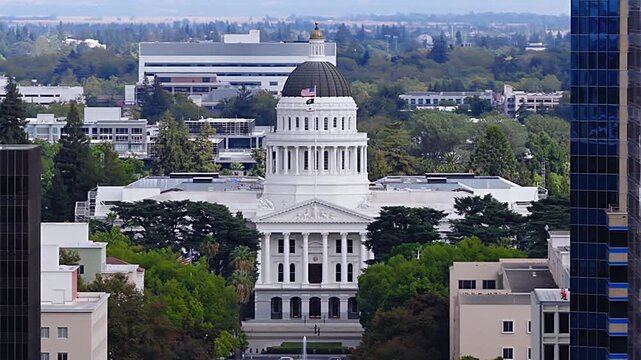 Aerial perspective reveals the historic California State Capitol building with its neo classical dome, portico, and lush park trees nestled between Sacramento downtown office blocks.
