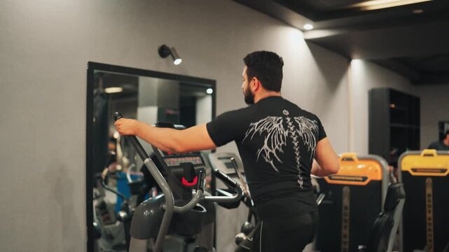 Three-quarter back view video shot of an athletic man training on an elliptical cross trainer in front of a wall mirror inside a modern commercial gym.
