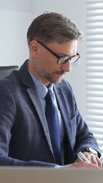 Mature entrepreneur in suit and tie wearing black glasses, is sitting at desk, writing notes and using calculator in modern, bright office. Audit and taxes in business