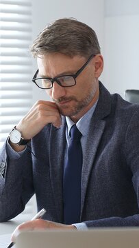 Adult businessman wearing blue suit and tail is concentrating and thinking while working at the desk in a bright, modern office, demonstrating professionalism and efficiency. Business people concept