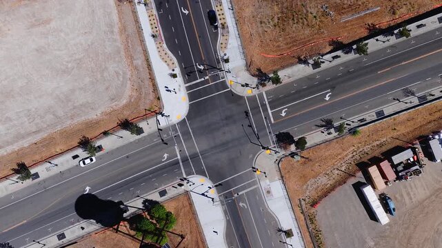 Aerial top down perspective of an empty four way asphalt road intersection with sidewalks, crosswalks, traffic arrows, and parked vehicles under bright daylight.
