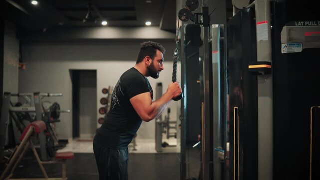 Side profile medium shot of a muscular bearded man performing triceps rope pushdowns on a cable machine for arm hypertrophy in a modern gym environment.