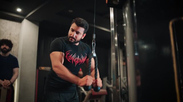 Medium close up video shot of a muscular bearded man looking away while completing a set of cable triceps pushdowns on a pulley machine in a modern gym with moody lighting.